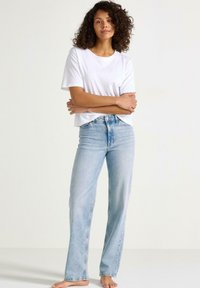 T-shirt bianco a maniche corte abbinato a jeans dritti blu sbiaditi. Cuffs arrotolati, vita alta e vestibilità rilassata. A piedi nudi, posa naturale.