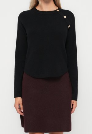 Femme portant un pull à manches longues noir avec trois boutons dorés sur l'épaule et une jupe bordeaux longueur genou.