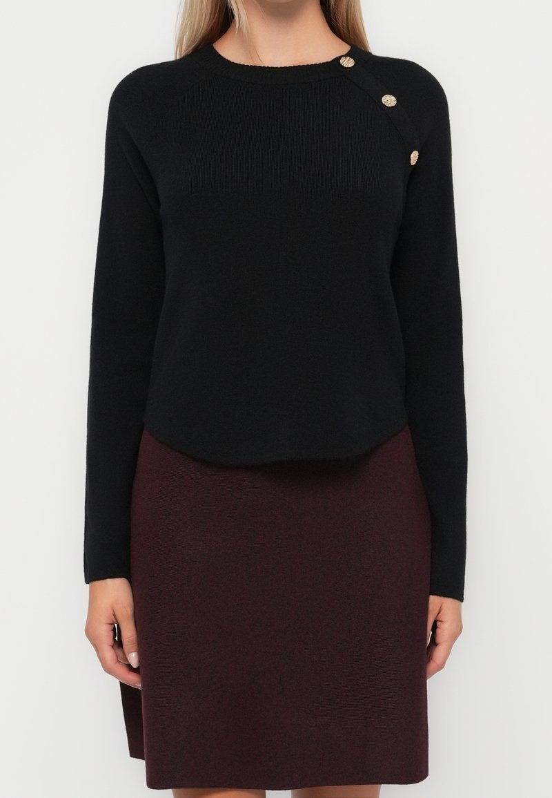 Femme portant un pull à manches longues noir avec trois boutons dorés sur l'épaule et une jupe bordeaux longueur genou.