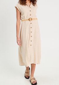 Robe midi en lin beige avec manches courtes, boutons à l'avant et deux poches, cintrée avec une ceinture à motifs. Portée avec des sandales à talons.