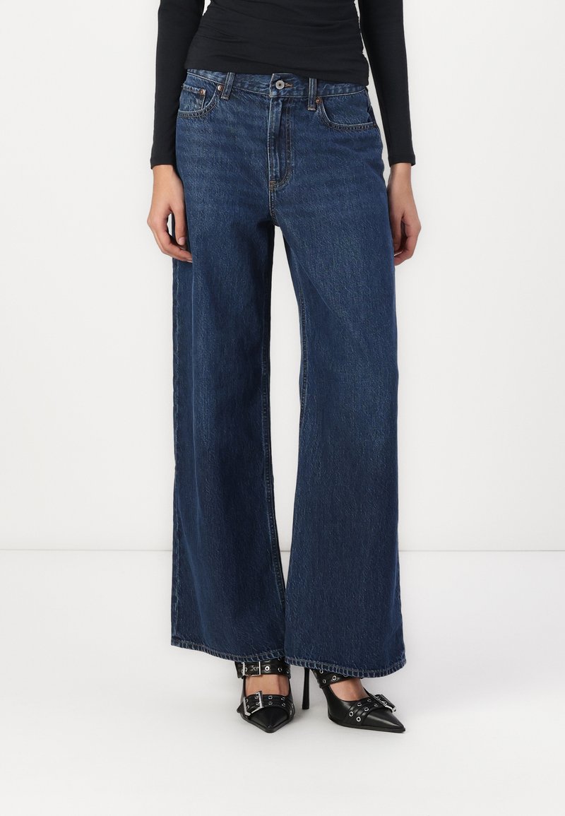 Jean en denim bleu foncé à taille haute et jambes larges, avec un design classique à cinq poches et des coutures contrastées. Assorti avec des chaussures noires à bouts pointus.