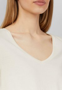Jasnekremowy top w serek wykonany z miękkiego materiału, z gładką teksturą i minimalnym przeszyciem. Dekolt ma czysty, subtelny brzeg.