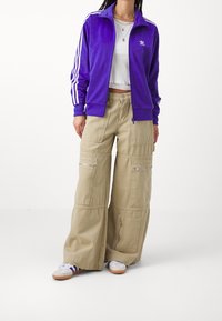 Lila velour zip-jacka med tre vita ränder på ärmarna, kombinerad med vida beige cargobyxor med flera fickor och blixtlås.