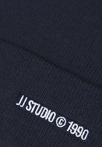 Navy gebreide beanie met ribbeltextuur. Bevat witte geborduurde tekst: "JJ STUDIO © 1990" nabij de onderkant.