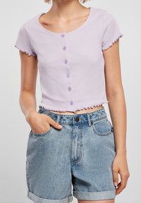 Levandulový top s krátkým rukávem, kulatým výstřihem, předními knoflíky a volánovými okraji. V kombinaci se světle modrými džíny s ohrnutými manžetami.