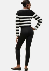 Gestreifter schwarz-weißer Strickpullover mit langen Ärmeln, kombiniert mit figurbetonten schwarzen Leggings und schwarzen Ballerinas. Bemerkenswerte Rippstruktur.