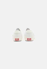 Bílé kožené slip-on tenisky s polstrovaným límcem, texturovanou patou a červeným logem Vans na zadní straně. Ploché gumové podrážky.