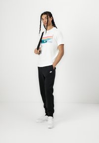 Balta īsām piedurknēm T-krekls ar krāsainu grafiku; melni sporta bikses; baltas kedas; cilvēks stāv pretī vienkāršai fona.