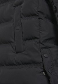 Tejido de chaqueta acolchada negra con secciones acolchadas y textura suave. Incluye un bolsillo con botón a presión y un detalle de herrajes en contraste.