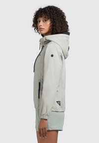 Lichtgrijze, waterdichte jas met een capuchon, elastische manchetten, zijzakken en een zwart accent op de kraag; heeft logo-details.