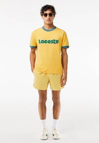 Żółta bawełniana koszulka z morską lamówką i logo "Lacoste". W połączeniu z pasującymi żółtymi szortami i białymi sneakersami. Luźny krój, krótkie rękawy.
