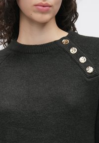 Tummanharmaa neulepaita, jossa on ribbineulottu pääntie ja neljä kiiltävää kultanappia olkapäällä. Pehmeä rakenne ja rento istuvuus.