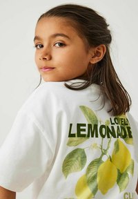 Witte katoenen T-shirt met groene tekst "Lovely Lemonade" en een grafische weergave van gele citroenen met groene bladeren op de achterkant. Korte mouwen.