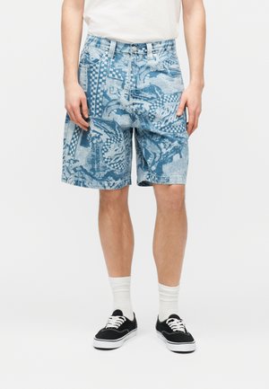 Άνδρας που φοράει μπλε τζιν σορτς με σχέδια, λευκές κάλτσες, μαύρα αθλητικά παπούτσια και ένα λευκό πουκάμισο που είναι κρυμμένο μέσα στο παντελόνι, στέκεται μπροστά σε απλό λευκό φόντο.