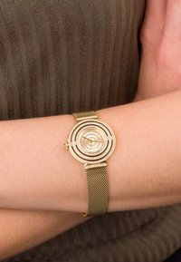 Zlatni mrežasti sat s okruglim skrivene uzorkom na brojčaniku ukrašenim rhinestones. Narukvica je finu tkanu metalnu mrežu.