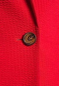 Button marrone a quattro fori con motivo a spirale etichettato "BOSS" su un tessuto rosso brillante a trama con un'asola verticale cucita.