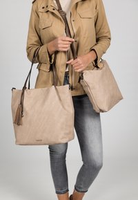 Two beige tote bags with brown leather straps, one large with a tassel accent and one smaller, held by a person in a khaki jacket.