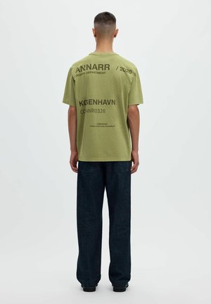 Mężczyzna stojący tyłem, ubrany w oliwkowo zielony T-shirt z czarnym napisem i ciemne, szerokie jeansy na jednolitym jasnym tle.