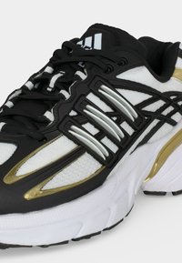 Chaussure de running noire et blanche avec des accents dorés, des lacets noirs et une semelle blanche épaisse, en mesh et matériaux synthétiques.