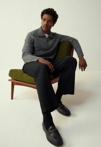 Harmaa vetoketjullinen neulepusero, jossa teksturoitu pinta, yhdistetty mustiin leveälahkeisiin housuihin ja mustiin loafer-kenkiin kultaisella yksityiskohdalla, istuu vihreällä tuolilla.