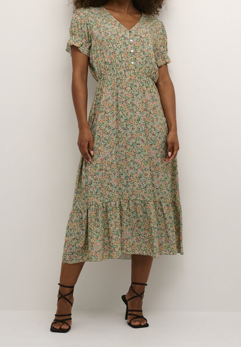 Femme portant une robe midi verte à imprimé floral, manches courtes, col en V boutonné, et des sandales noires à talons hauts avec des brides, debout contre un mur uni.