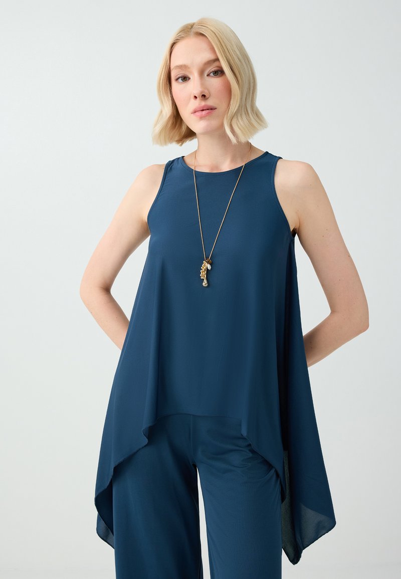 Femme aux cheveux blonds courts portant une tunique sans manches bleu marine, un pantalon assorti et un long collier doré, debout devant un fond uni.