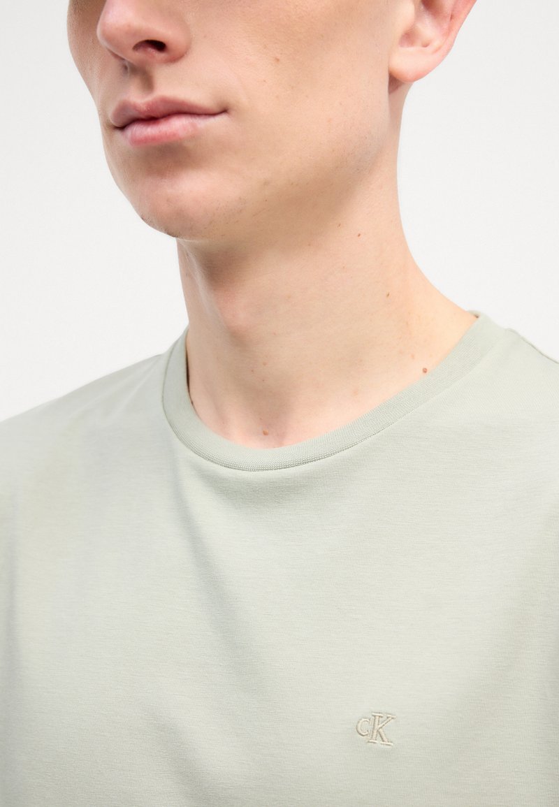 Light green crew neck t-shirt made of soft cotton, features a subtle embroidered logo on the chest in a matching color. Smooth texture.