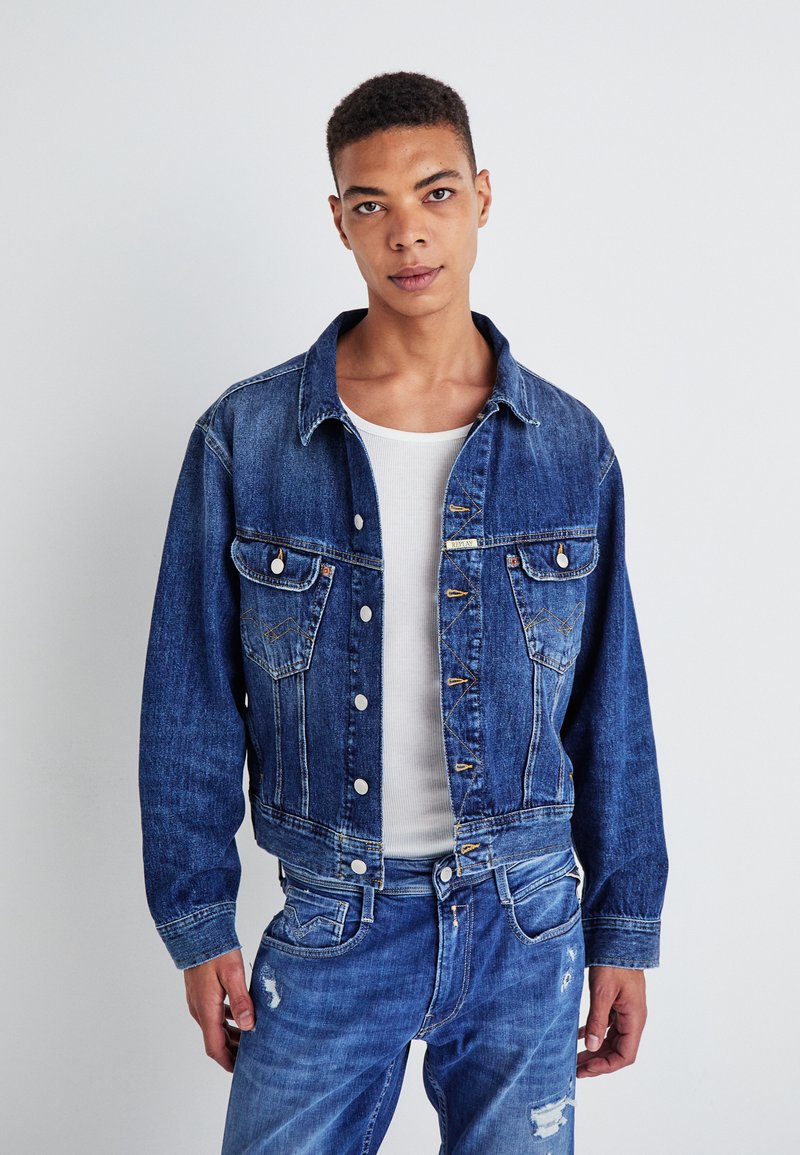 Denim jacket, blue, with two chest pockets, button closure, and a cropped design. Worn over a white ribbed shirt and matched with blue jeans.