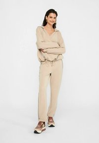 Beżowa bluza z kapturem ze sznurkami, w zestawie z dopasowanymi zwężanymi spodniami. Oba wykonane z miękkiego materiału; posiadają boczne akcenty. Brązowe buty sportowe dopełniają wygląd.