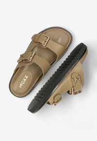 Tanové skluzavky vyrobené z hladkého materiálu, s dvěma nastavitelnými popruhy se zlatými přezkami a texturovanou černou podrážkou.