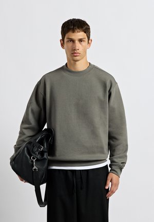 Ung mand iført en grå sweatshirt og sorte bukser, der holder en stor sort lædertaske med skulderrem.