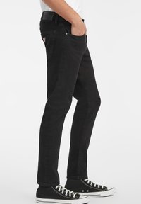 Svart jeans med slim fit-design, fem lommer, metallbeslag og en subtil tekstur. Kombinert med svarte høye sneakers.