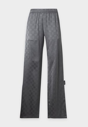 Mujer de pie con camisa gris suelta con estampado y pantalones, sosteniendo un bolso negro al hombro, con zapatos negros de punta abierta.