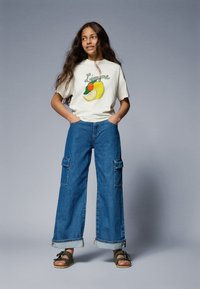 Cream graphic t-shirt with lemon design, paired with wide-leg blue denim cargo pants featuring side pockets and turned-up cuffs. Brown sandals.