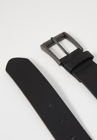 Ceinture en cuir noir avec une finition mate, dotée d'une boucle métallique à texture brossée. Comprend plusieurs trous d'ajustement pour le dimensionnement.