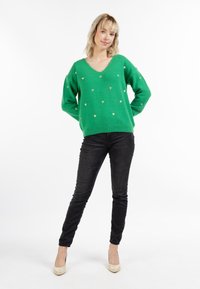 Maglione verde a V con motivi a cuore dorati, abbinato a jeans skinny neri e scarpe con tacco beige. Il tessuto sembra morbido.