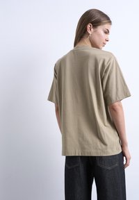 Νεαρό άτομο με ίσια μαλλιά που φοράει ασημί σκουλαρίκια κρίκους, μία μπεζ υπερμεγέθη κοντομάνικη μπλούζα και σκουρόχρωμα τζιν, κοιτάζοντας πλευρικά.