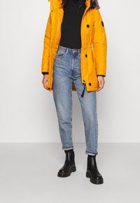 Personne portant un manteau d'hiver jaune vif avec capuche en fourrure, un col roulé noir, un jean bleu clair et des bottines noires, debout sur un fond uni.