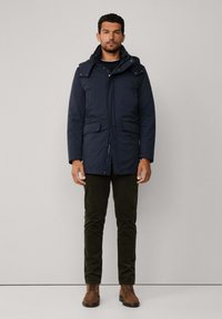 Mężczyzna w granatowej zimowej kurtce z kapturem, ciemnych spodniach i brązowych zamszowych butach na sznurowadła, stojący na jednolitym jasnym tle.