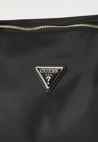 Sac en nylon noir avec une fermeture éclair dorée et un badge logo noir triangulaire avec le texte "GUESS U.S.A." et un motif de point d'interrogation.