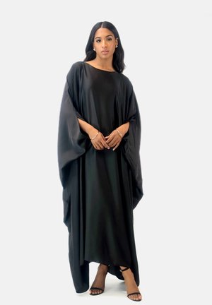 Black, oversized dress with a flowing design, made of smooth fabric. Features wide sleeves and a rounded neckline. Worn with black heeled sandals.