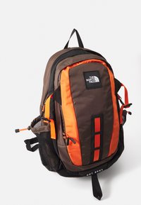 Barna és narancssárga North Face hátizsák, több rekesszel, hálós oldalsó zsebekkel és állítható pántokkal, fehér háttér előtt.