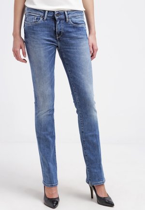 Schmal geschnittene blaue Jeans, getragen von einer Person, kombiniert mit schwarzen High-Heel-Schuhen und einem weißen Oberteil, stehend vor einem schlichten Hintergrund.