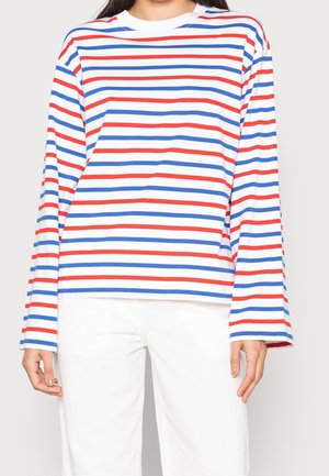 Personne portant un haut blanc à manches longues avec des rayures horizontales rouges et bleues, associé à un pantalon blanc, présenté des épaules aux hanches.