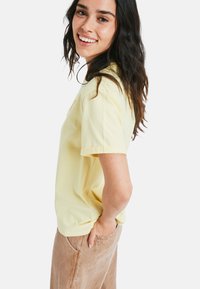 Gelbes Baumwoll-T-Shirt mit kurzen Ärmeln, rundem Ausschnitt und lockerer Passform, kombiniert mit hellbraunen Hosen. Einfaches, einfarbiges Design.