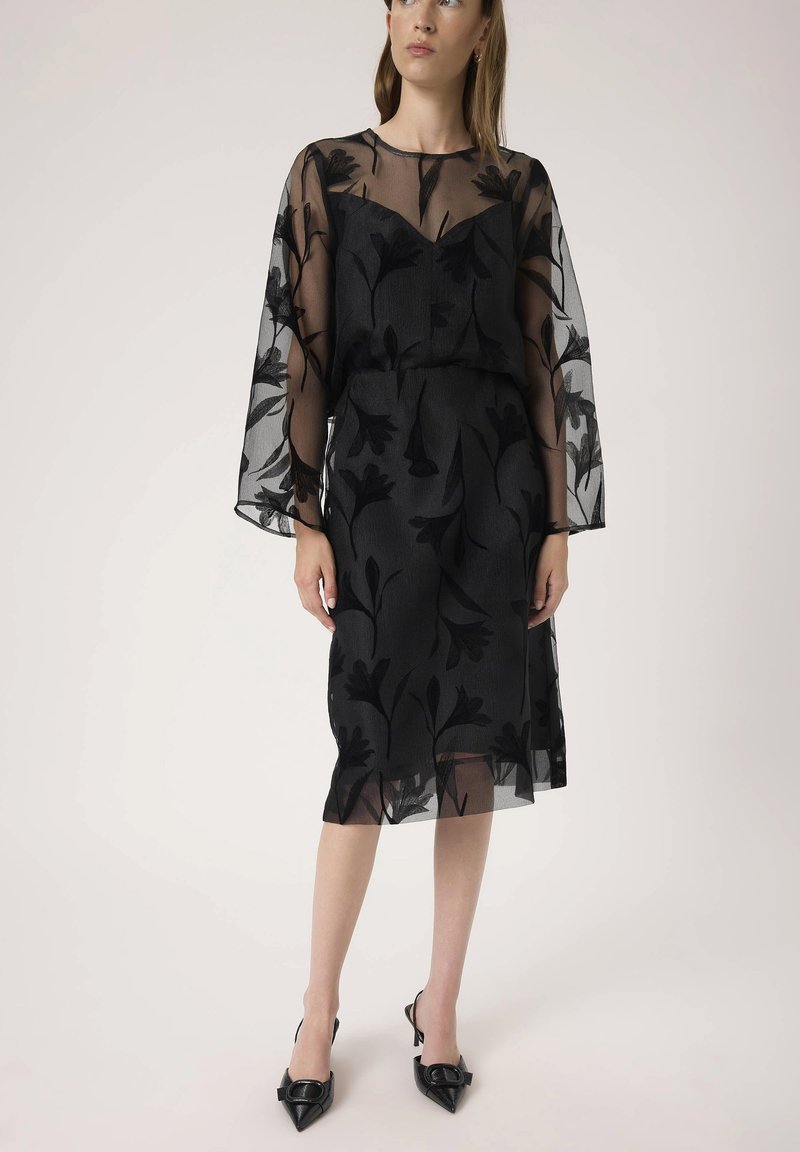 Rochie neagră cu mâneci lungi din material transparent, broderie florală și talie ajustată. Fusta se termină puțin sub genunchi, fiind completată de tocuri negre ascuțite.