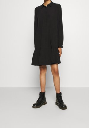 Woman wearing a long-sleeve black collared dress with a tiered skirt, paired with black lace-up combat boots, standing against a plain background.