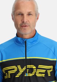 Maglia sportiva zip-up blu e nera con strisce gialle e un logo, colletto alto e tessuto liscio e leggero.
