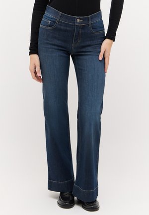 Person iført mørkeblå bootcut-jeans, sorte sko og en sort langærmet top, stående mod en ensfarvet lys baggrund.