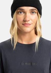 Svart ribbet lue på lys blonde hår. En mørk grå Quiksilver hettegenser har rund hals og brodert logo på brystet.